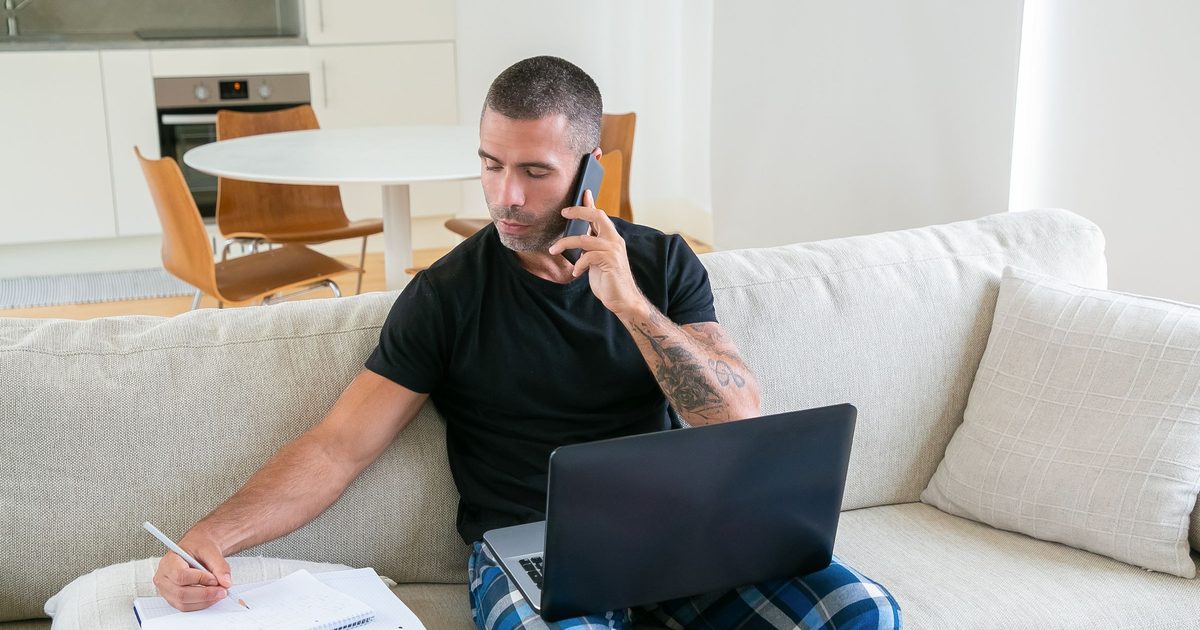 Man sitting on a couch taking notes on a notepad while on a phone call, preparing for a first recruiter conversation